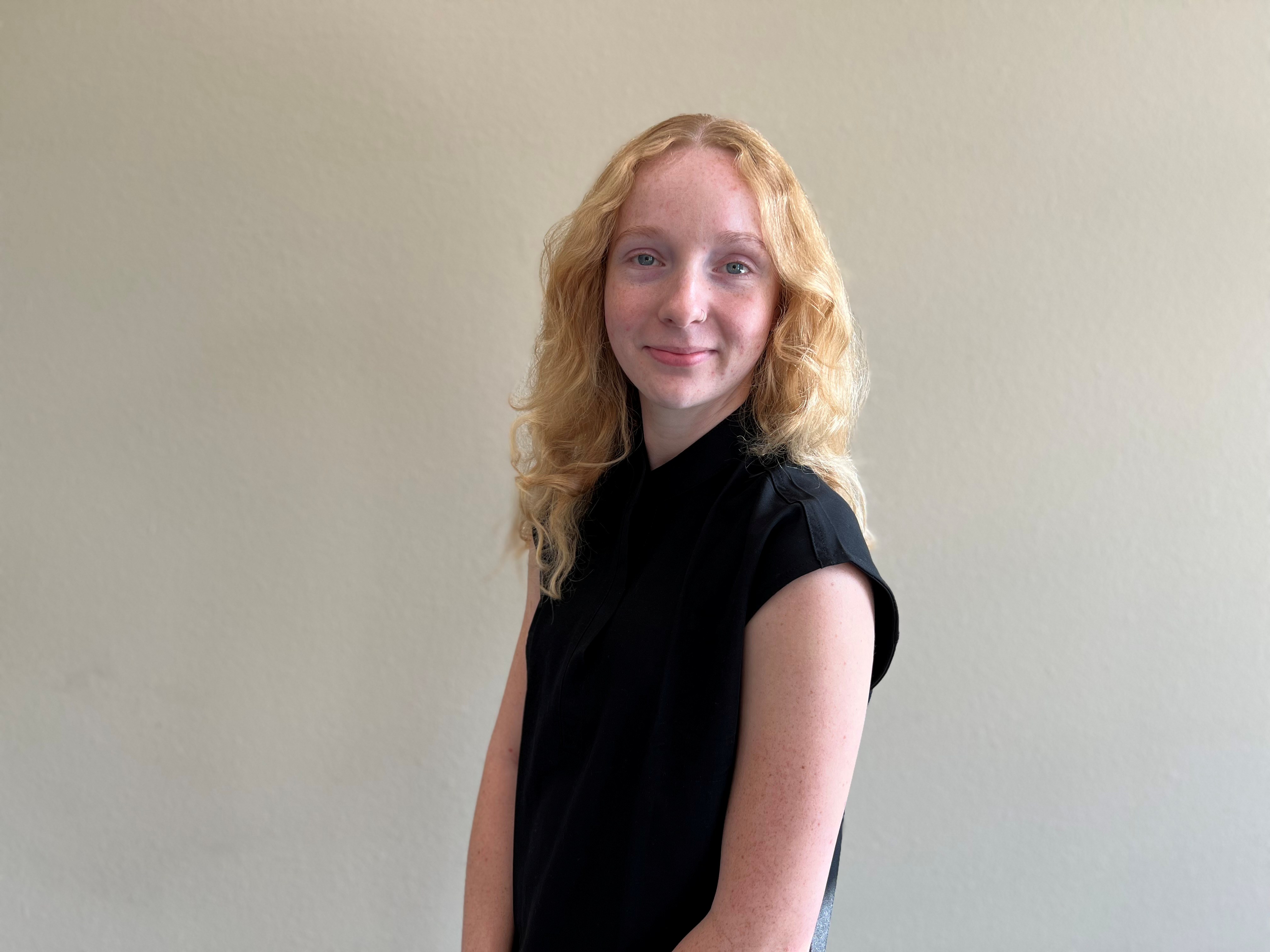 A person smiling while standing up against a blank wall